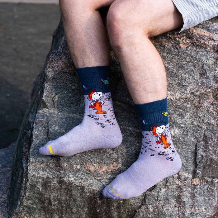 A person sitting on a large rock wearing light grey crew socks with a dark navy cuff and a cartoon character printed on the lower half. The character is wearing an orange space suit, with small stars and a small yellow-green planet detail.