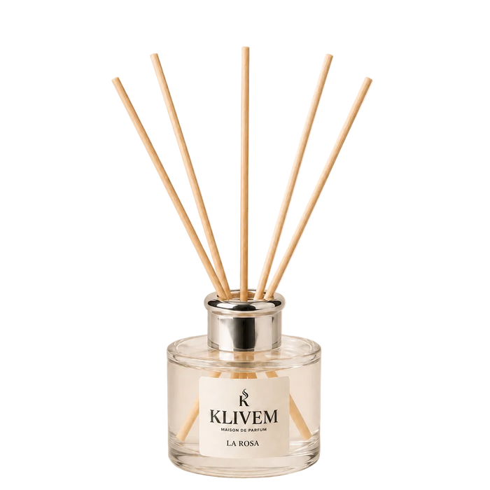 A reed diffuser bottle with a clear glass base, silver metal collar, and six natural wood reeds, featuring a white label with the brand name KLIVEM and scent name LA ROSA, set against a solid pale beige background.
