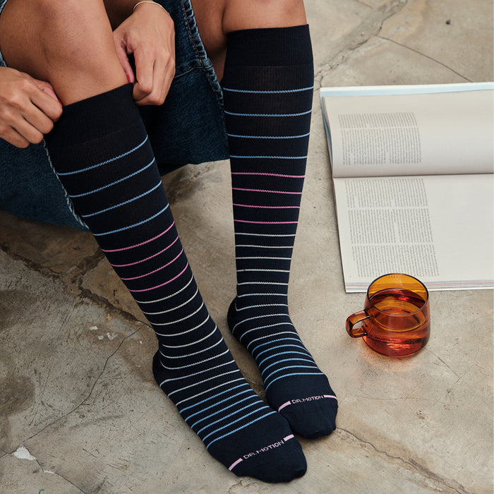 Knee-high compression socks with multi-color pinstripes in blue, pink, and gray, worn by a person sitting on a concrete surfa